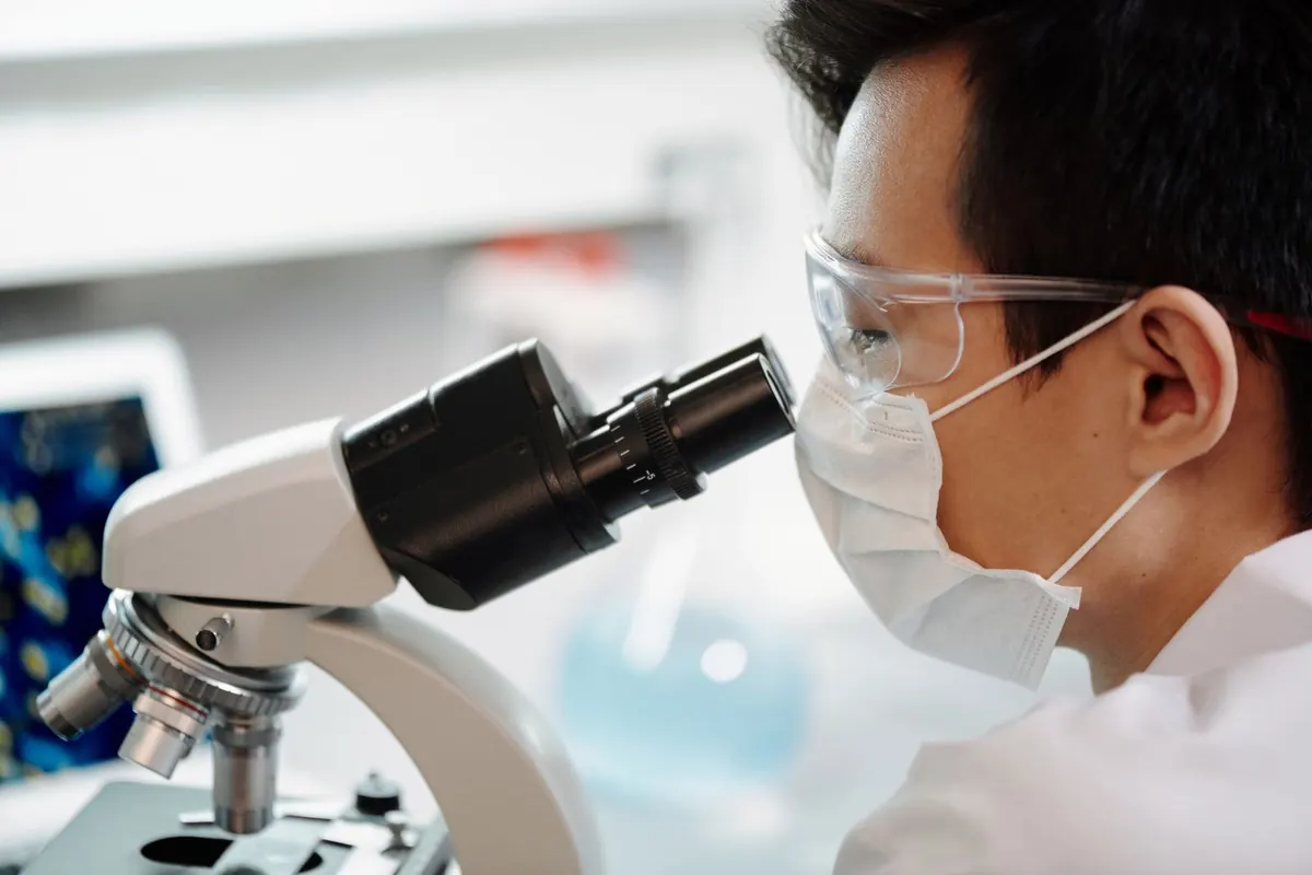 Scientist in goggles and a face mask looking through a microscope in a bright lab