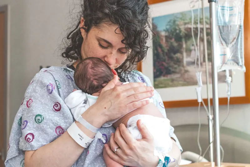 Mother in a hospital gown gently kissing her sleeping newborn held against her chest