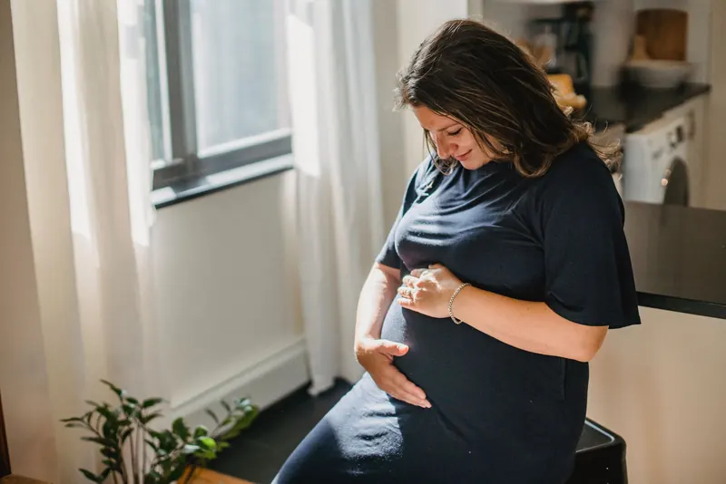 Pregnant woman sitting by a window in soft daylight with her hands resting on her belly