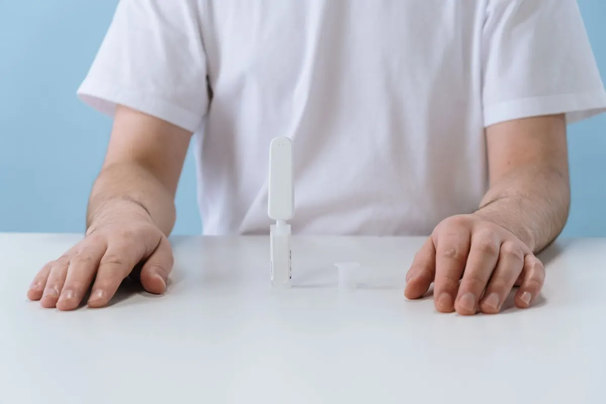 Person in a white T-shirt seated at a white table with a small at-home test device in front of them