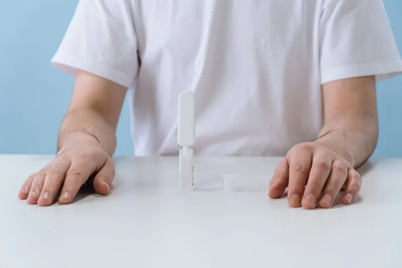 Person in a white T-shirt seated at a white table with a small at-home test device in front of them
