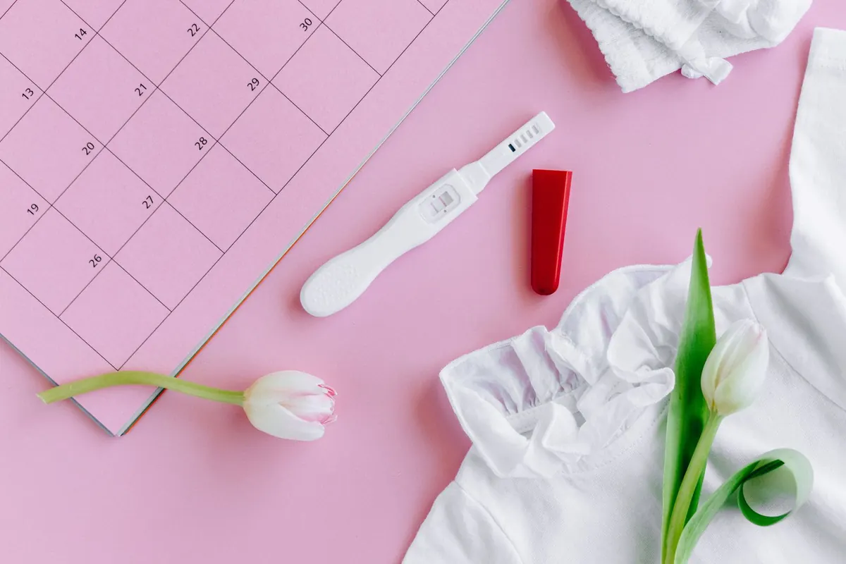 Ovulation test stick on a pink surface next to a calendar page, white tulips and a white top