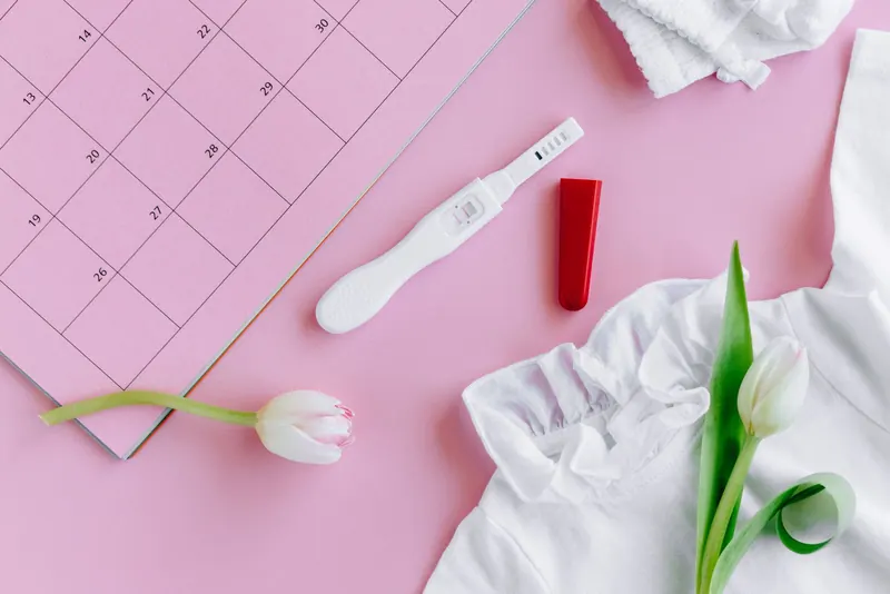 Ovulation test stick on a pink surface next to a calendar page, white tulips and a white top