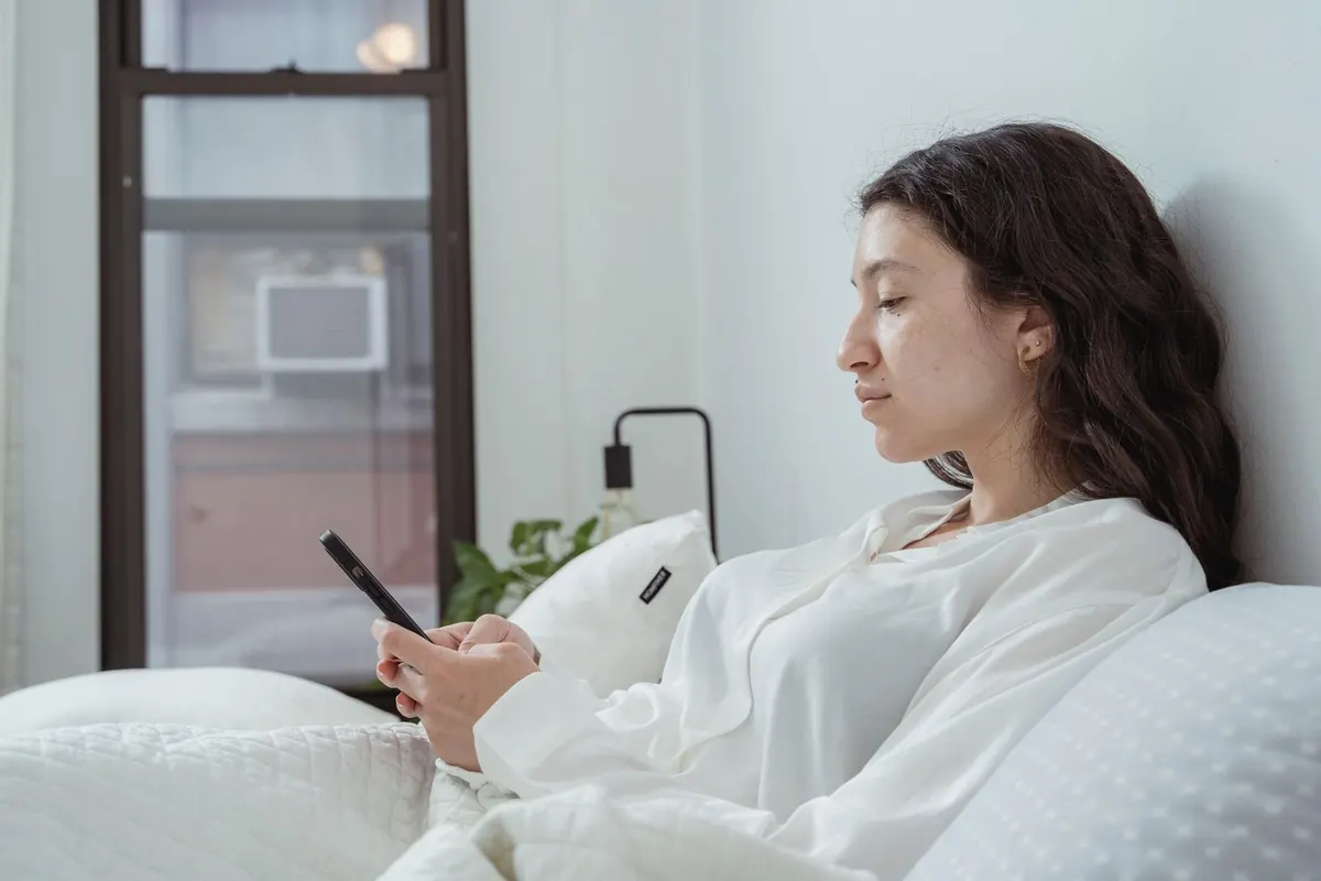 Woman in white sleepwear sitting up in bed checking her phone in soft morning light