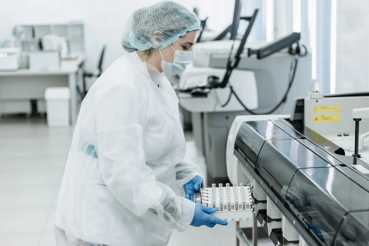Lab technician in a hair cap and gloves loading a rack of sample tubes into an analyzer in a bright lab