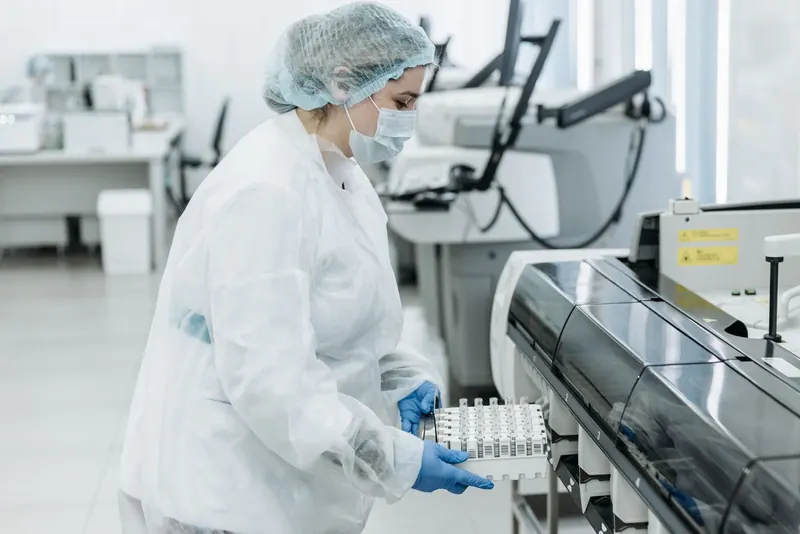 Lab technician in a hair cap and gloves loading a rack of sample tubes into an analyzer in a bright lab