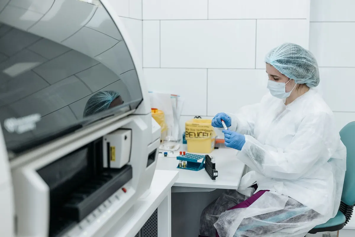 Lab technician in protective gown and gloves handling sample tubes beside an analyzer in a bright lab