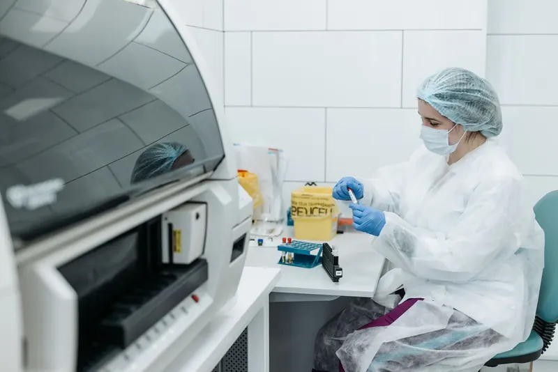 Lab technician in protective gown and gloves handling sample tubes beside an analyzer in a bright lab