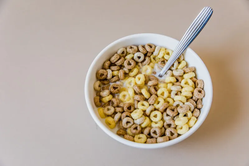 White bowl of ring-shaped breakfast cereal with milk and a spoon on a light table
