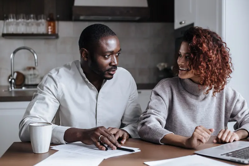 Couple sitting at a kitchen table with papers and a laptop, talking through plans together